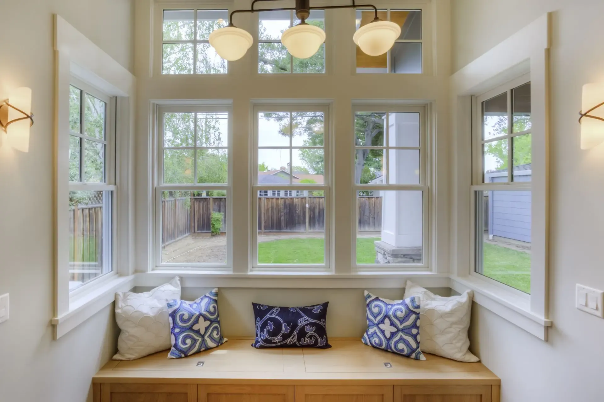 Bright sunroom with window seat featuring blue and white patterned pillows, three-pendant light fixture above, multiple large windows overlooking backyard and patio area