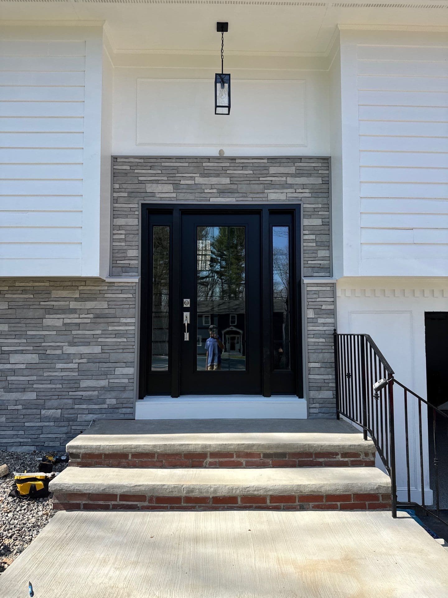 Modern home entryway with black double-glass doors flanked by sidelights, gray stone veneer surround, brick and concrete steps, metal railing, and hanging pendant light above the entrance
