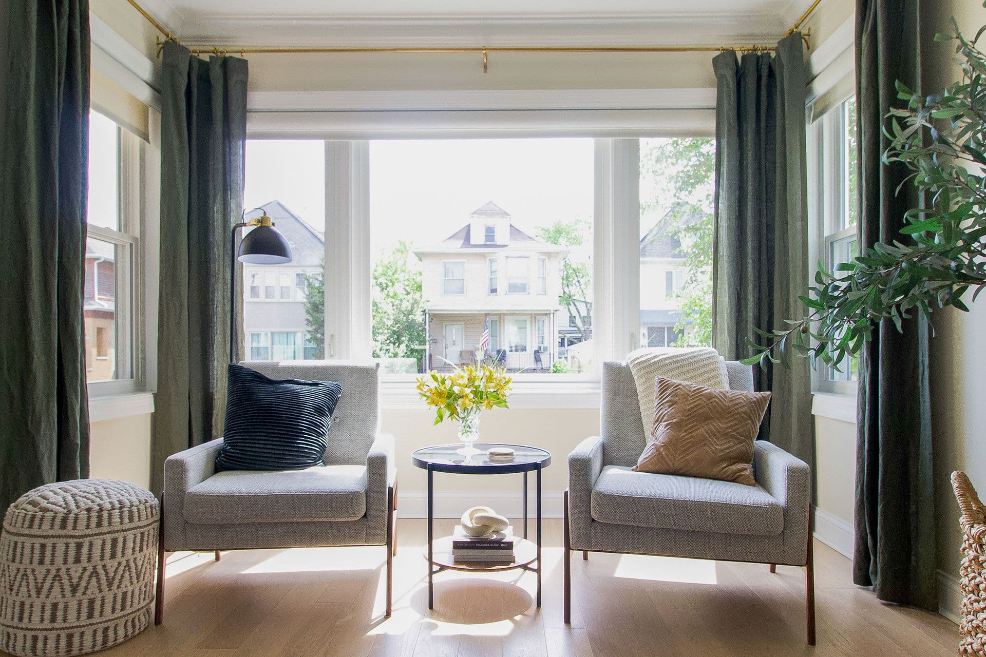 Bright sunroom with two gray upholstered armchairs facing a round side table with flowers, flanked by large windows overlooking a residential street with historic homes