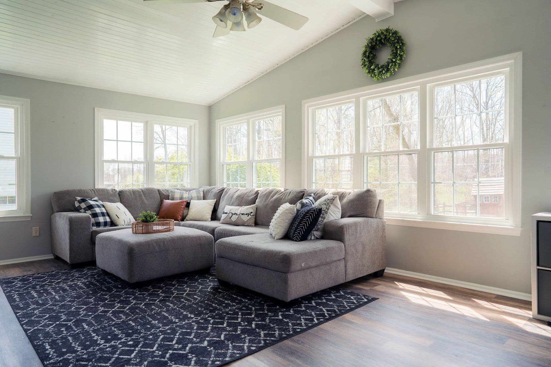 Bright living room with gray sectional sofa adorned with decorative pillows, positioned on a patterned blue rug, large windows overlooking outdoor landscape, and a green wreath on the wall
