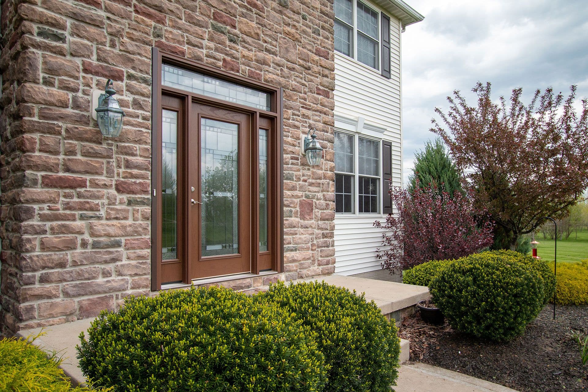 Brick home exterior with brown double-glass doors flanked by lanterns, concrete entryway, and landscaped shrubs in foreground