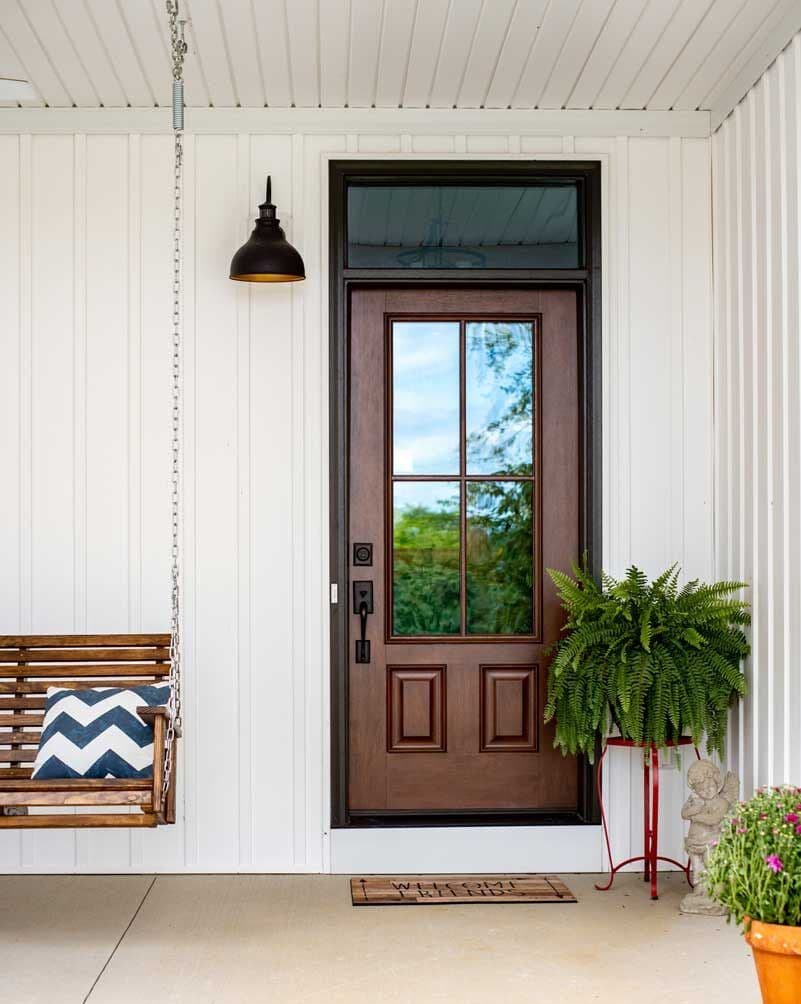 Front porch with brown wooden door, black pendant light, hanging swing with chevron pillow, fern in red planter, and rural landscape visible through door glass