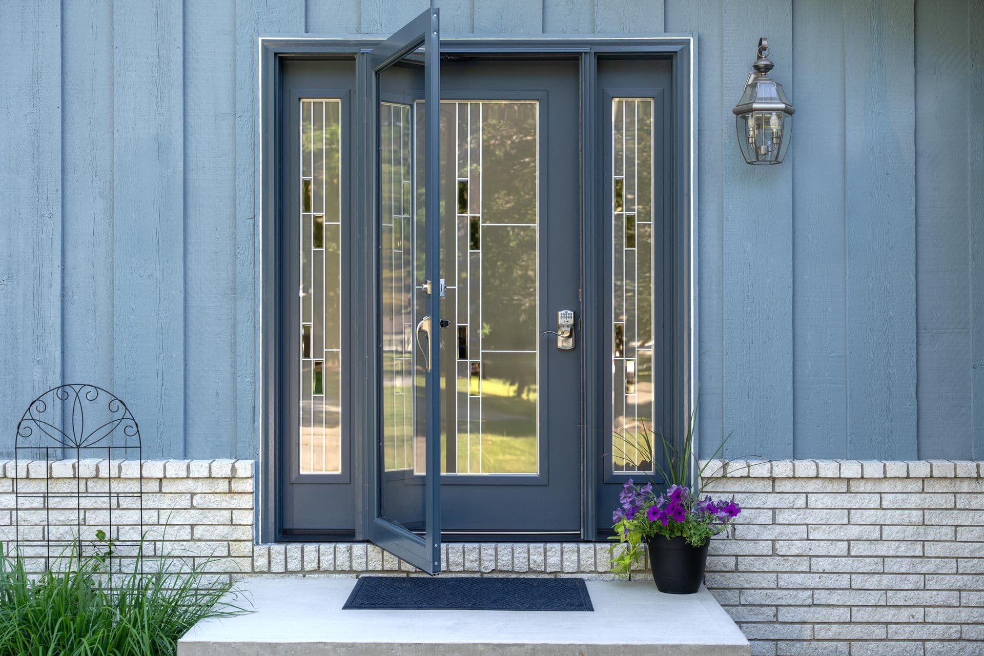 Modern home entrance with dark blue double doors featuring sidelights, flanked by white brick walls, welcome mat, purple flowering planter, and exterior lantern sconce