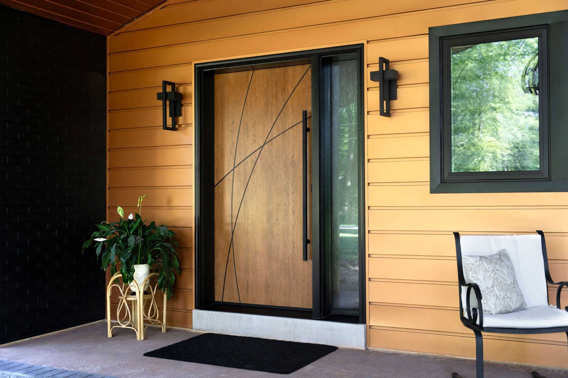 Modern home entrance with warm wood siding, black-framed door with geometric pattern, side window, two decorative wall sconces, potted plant, black entry mat, and seating area with cushion